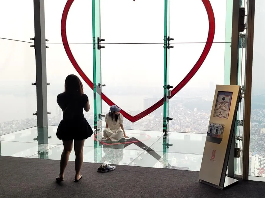 Visitor sitting on a glass observation floor in front of a large red heart sculpture, the city far below
