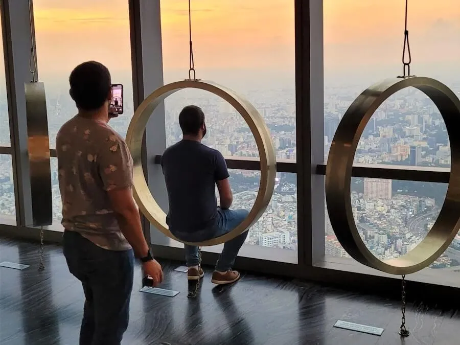 Two visitors at the floor-to-ceiling windows of a high observation deck at sunset, one seated in a hanging ring swing